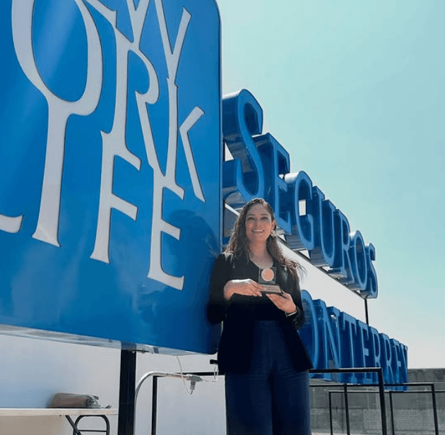 Asesora de Godshall Consultores con un reconocimiento frente al letrero de Seguros Monterrey New York Life.